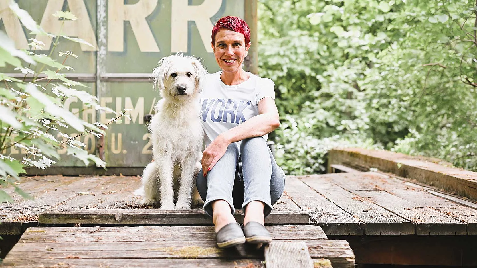 Nadine Pötz mit Hund in der Natur Frau sitzt mit einem Hund vor einer grünen, bewachsenen Kulisse.