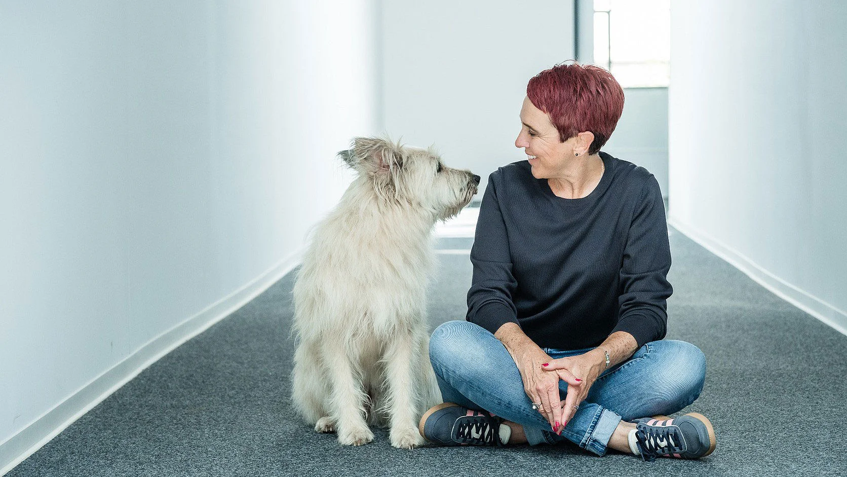 Frau sitzt mit einem weißen Hund auf dem Boden eines hellen Raumes.